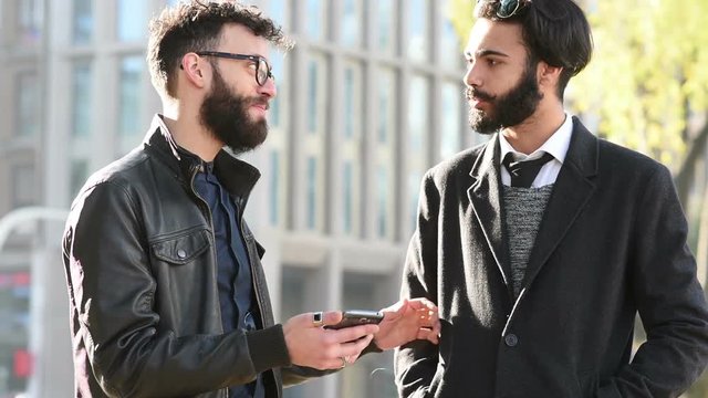 Half length of two young handsome caucasian bearded brown hair businessmen walking in the city, talking to each other with smart phone handhold- business, technology, work concept