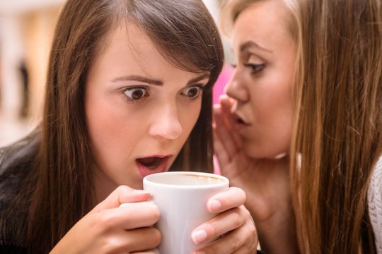 Woman whispering in her friends ears while having coffee