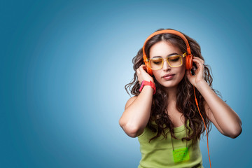  Girl in headphones and glasses looking down and listening to music, enjoying music.Closeup portrait girl on blue background. Positive human emotion facial expression feeling