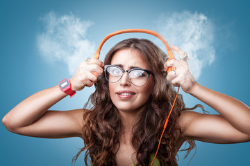 Surprised impressed girl with long curly hair in headphones listening to music, blowing white smoke coming out of ears.Closeup portrait girl on blue background. Human emotion facial expression feeling