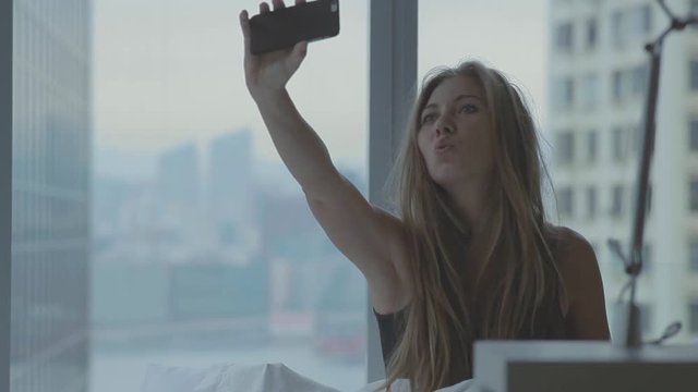 Young blonde woman with long hair making selfie with her smart phone in the bed in the morning. Handheld shot. Hong Kong, China.
