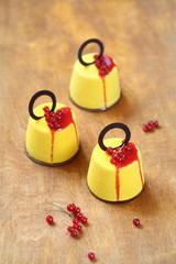 Contemporary Mousse Cakes covered with yellow velvet spray, decorated with red berries topping, red currants and chocolate rings, on a wooden table.