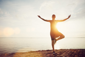 Obraz premium Young woman standing in yoga pose on the beach and meeting sunrise. Intentional sun glare, lens flare effect