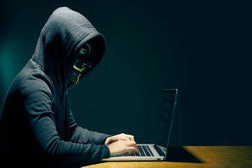 Side view of person wearing hooded top and Guy Fawkes face mask using laptop