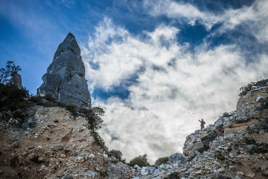Low angle view of young man standing on cliff pointing at cone shape rock formation, Ogliastra, Sardinia, Italy