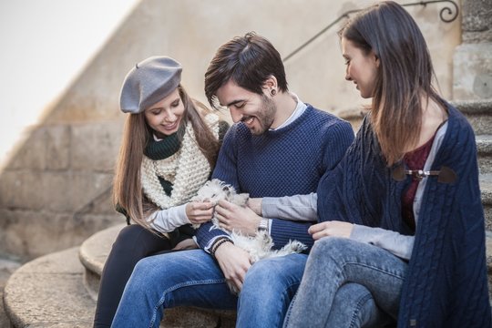 Friends sitting side by side on steps holding fluffy dog looking down smiling
