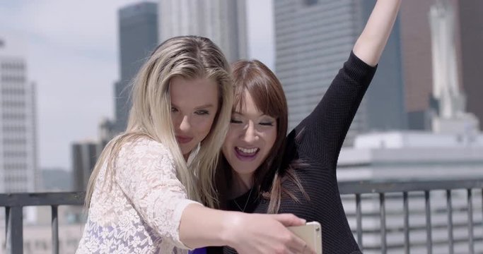 Close up of two young women posing for cellphone camera, smiling and laughing, in front of the Downtown Los Angeles skyline.  Slow motion, recorded in 4K at 60fps.
