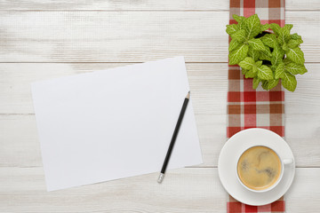 Cup of coffee and indoor plant are on a checkered tablecloth with white paper, pencil next to them.