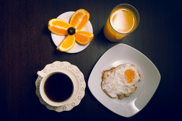 Breakfast on dark wood surface including coffee, bread, orange juice, fried egg and slices of oranges placed upon small white plate