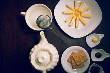 Plates of buiscuits and cheeses organized in star formation on dark wooden surface next to tea kettle, small plates with jam plus butter, shot from above angle