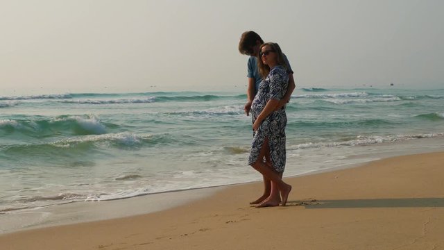 Caucasian couple hugs at the seashore. Man caressing his pregnant wife's belly. Splendid seascape.