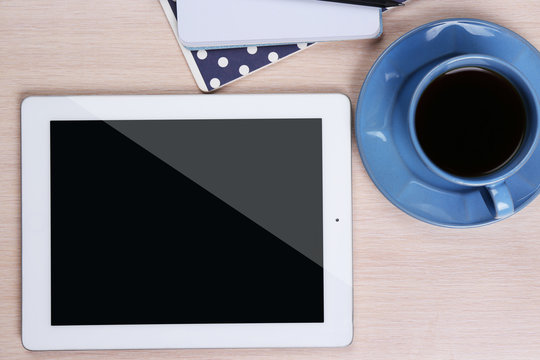 Tablet with cup of coffee and notepad on wooden table