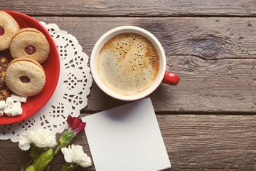 Cup of coffee with cookies, flowers and blank piece of paper on wooden background