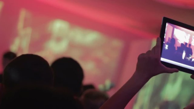 Male fan enjoying concert and filming performance on tablet, crowd in night club