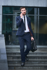 A businessman walks down the stairs and talking on mobile phone holding a briefcase against the building with a glass facade © amixstudio