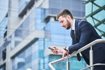 Businessman looking at phone in hand leaning on the railing against the building with a glass facade