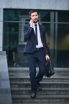 A businessman walks down the stairs and talking on mobile phone holding a briefcase against the building with a glass facade