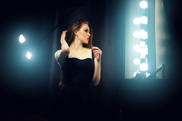 Beautiful sexy young caucasian woman in black dress, looking into makeup mirror at herself. Backstage dramatic shot.