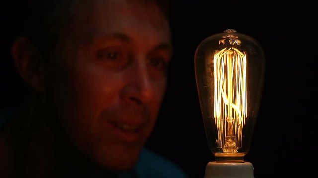 Close up shot of a man fascinated by an antique style filament bulb with his face barely lit by the soft warm yellow light of the retro light source.