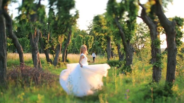 Beautiful young blonde bride running in the green spring forest lit by sunbeams.  Handsome groom on background