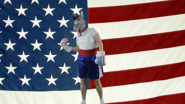 Woman wearing Democrat donkey mask wearing boxing shorts and gloves punch self in head and face against American Flag.