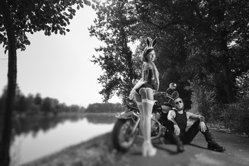Sexy fashion female in bunny costume and biker with long beard by motorcycle. Man sitting on the ground woman standing next to him. Summer day. Tilt shift lens blur effect. Black and white
