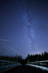 Un ciel nocturne plein d& 39 étoiles et une voie lactée visible avec un pont au premier plan. Route menant à la forêt sombre.