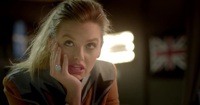 Close up of beautiful young blonde woman sitting in urban bar lounge and looking straight to camera, then mouthing "when?" or "what?" Hand held, slow motion, recorded in 4K at 60fps.