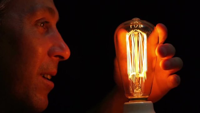 Close up side view shot of a man fascinated by an antique style filament bulb touches it and taps the glass bulb with his finger.