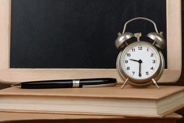 silver wind-up alarm clock near wooden boards for writing and book with pen on a wooden table