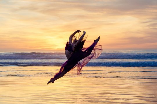 Young female dancer wearing chiffon dress, dancing, in mid air, on beach at sunset, San Diego, California, USA