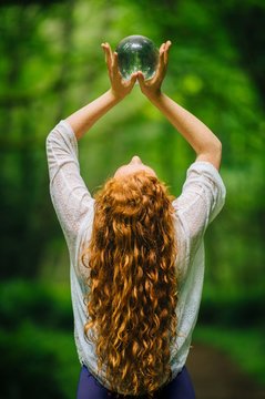 Rear view of young woman with long red hair holding up crystal ball in forest