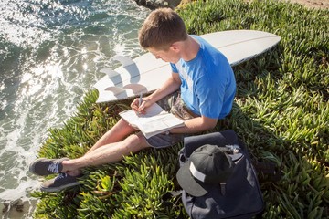 Young man sitting by cliff edge, writing in book, elevated view