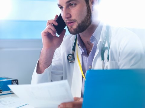 Junior Doctor Using Smartphone Whilst Reviewing Medical Records In Clinic