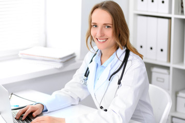 Beautiful young smiling female doctor sitting at the table near window in hospital