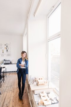 Full length view of young woman in office leaning against wall arms crossed looking away out of window