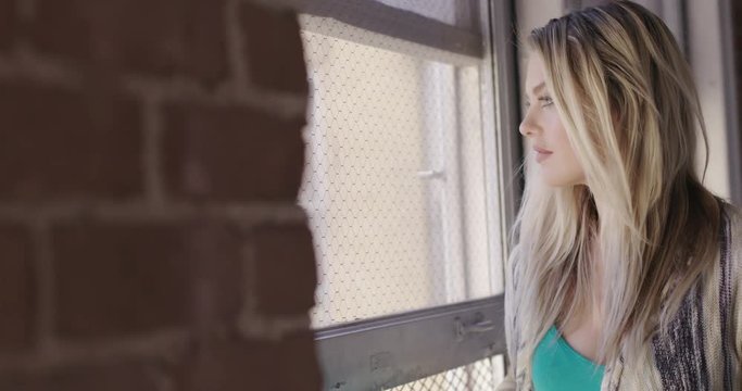 Close up of beautiful young blonde woman looking out of a window in an urban hallway with brick walls. Hand held, slow motion, recorded in 4K at 60fps.
