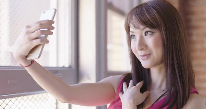Close up of attractive young Asian woman taking selfies with a cellphone next to a window in an urban setting.  Low angle camera, slow motion, recorded in 4K at 60fps.