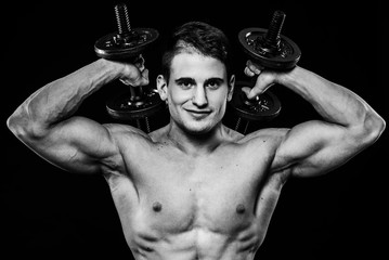 Handsome power athletic man training pumping up muscles with dumbbells in a gym. Barbells behind the head. Fitness muscular body isolated on black background. Looking to camera  smiling. Front view