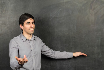Portrait of young happy smiling teacher man standing near chalkboard background and showing...