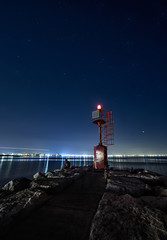lighthouse and rocks night seascape. City lights after sunset. R