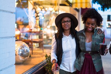 Lesbian couple walking along street, arms around each other, carrying takeaway drinks
