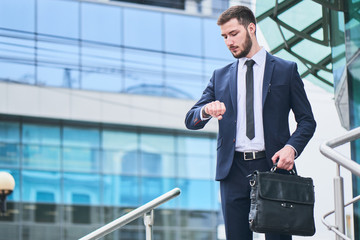 Businessman stands holding a briefcase and look at the clock on the background of the building with...