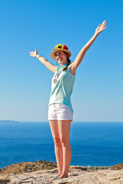 Beautiful girl with open arms in the air like happy on a summer day with the ocean behind her and wearing shorts and a straw hat. 