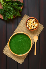Fresh homemade cream of spinach soup in wooden bowl, homemade croutons, wooden spoon and spinach leaves on the side, photographed overhead on dark wood with natural light
