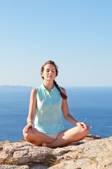 Fototapeta na wymiar Beautiful young woman meditating outside on the top of the mountain with the blue sea behind her.