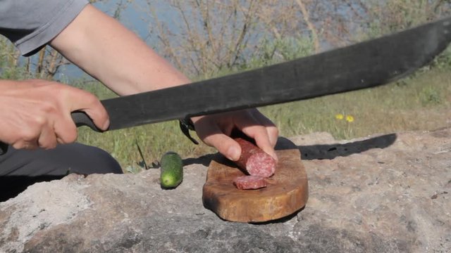 Man slices salami on the stone using a machete

