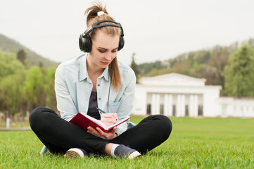 Fototapeta premium Young beautiful female sitting and writing outside with earphone