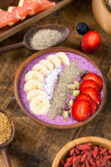 Fototapeta na wymiarAcai berry smoothie bowl topped with chia seeds, coconut flakes, banana, strawberries and kiwi with variety of superfoods on rustic wooden table. Healthy breakfast. Selective focus