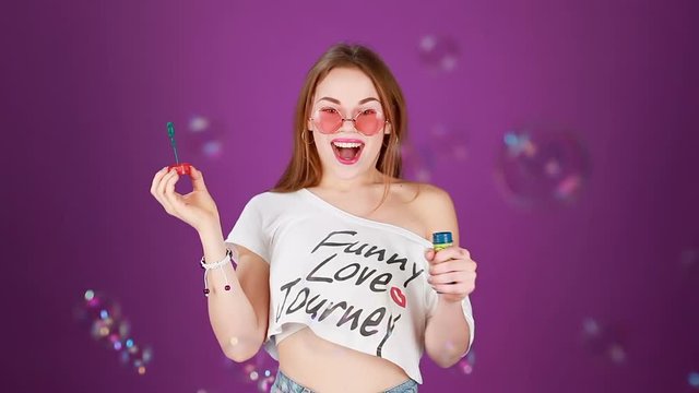 Funny fashion girl blowing soap bubbles on purple background. Hipster teenager female in pink round glasses.